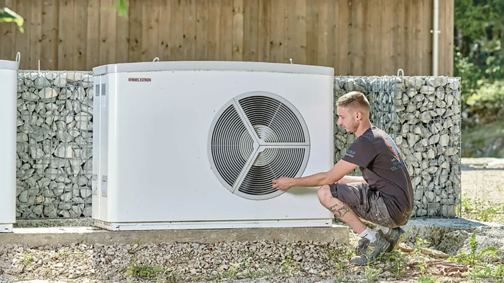 Technicien en combinaison de travail effectuant la maintenance d'une pompe à chaleur air-eau extérieure blanche, installée sur dalle béton avec écrans acoustiques en gabions