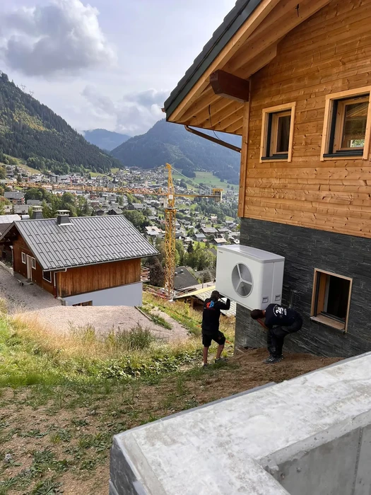 Deux techniciens installent une pompe à chaleur air-eau sur façade extérieure d'un chalet moderne en bois et ardoise